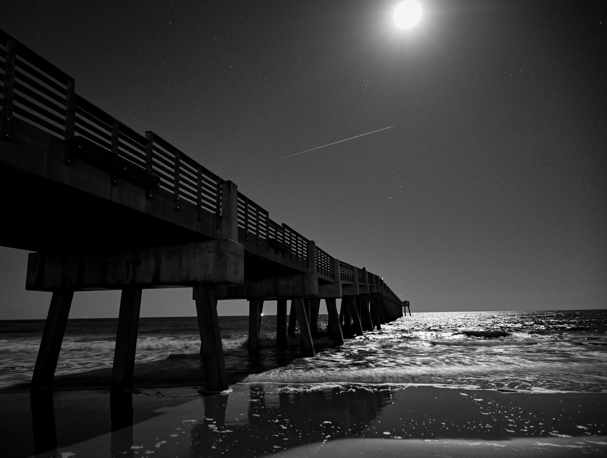 The Jacksonville Beach pier under a full beaver moon with the light trail of an airplane tracing across the night sky.