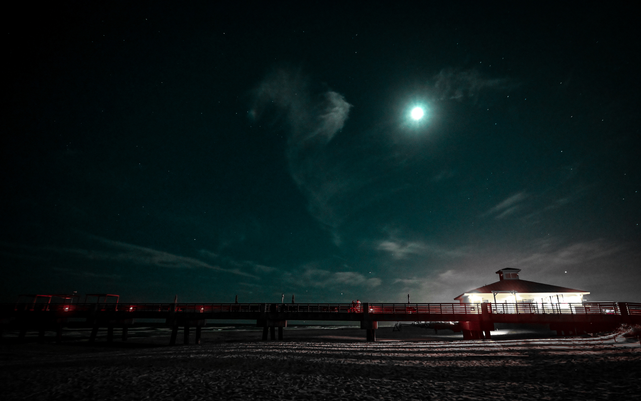 The Jacksonville Beach pier under a cyan moonlit night with wispy clouds.