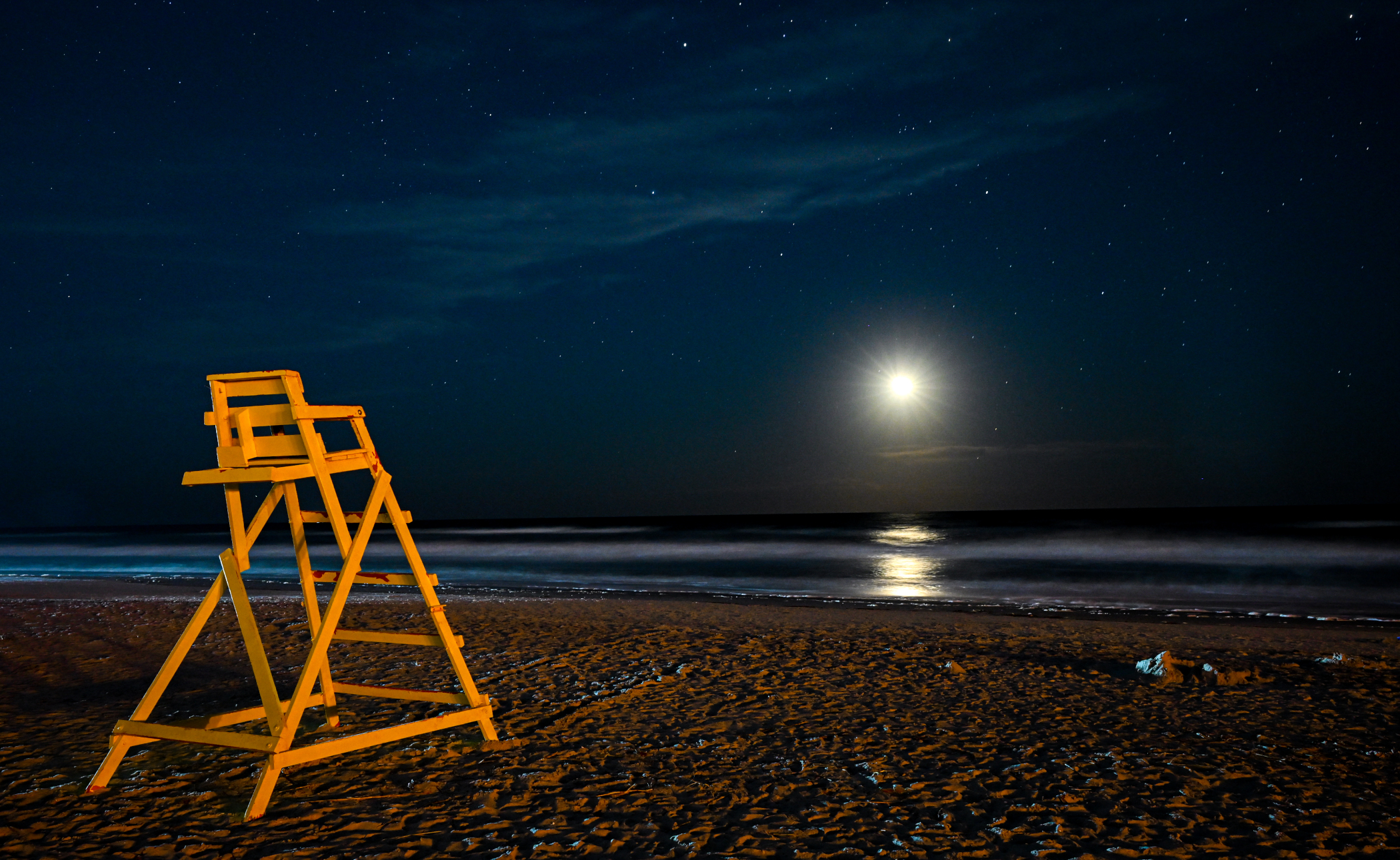 A yellow lifeguard chair on Jacksonville Beach Florida under a starry dark blue night sky illuminated by a dazzling full moon.